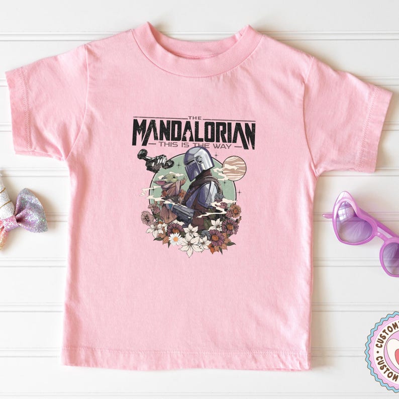 Może przedstawiać: R&oacute;żowa koszulka z nadrukiem The Mandalorian i Grogu, otoczonych kwiatami, oraz napisem "THE MANDALORIAN - THIS IS THE WAY" w kolorze czarnym. Projekt znajduje się z przodu koszulki z kr&oacute;tkim rękawem.