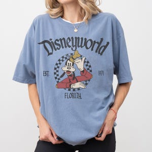 May include: A light blue t-shirt featuring a graphic of Goofy from Disney World. The graphic is in a circular design with the words "Disneyworld", "Florida", and "Est. 1971". The shirt has short sleeves and a crew neck.