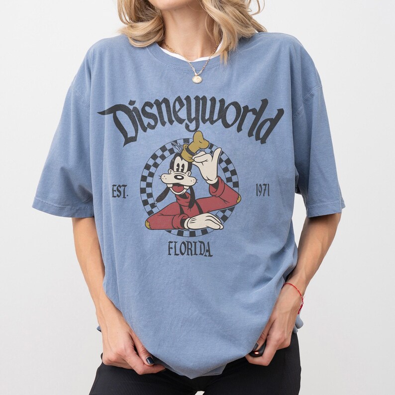 May include: A light blue t-shirt featuring a graphic of Goofy from Disney World. The graphic is in a circular design with the words "Disneyworld", "Florida", and "Est. 1971". The shirt has short sleeves and a crew neck.