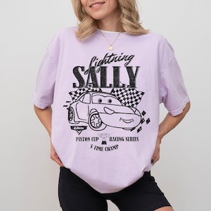 May include: A lilac t-shirt with a black and white graphic of a cartoon car and the text "Lightning Sally". The design includes a checkered flag and the words "Piston Cup Racing Series 5 Time Champ".