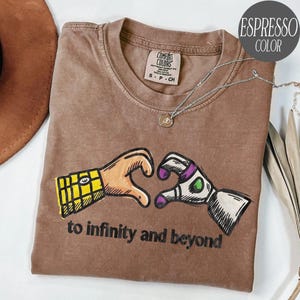 May include: An espresso-colored t-shirt featuring a graphic of two hands forming a heart shape, one with a yellow plaid pattern and the other with white, purple, and green details. The text "to infinity and beyond" is printed in black below.