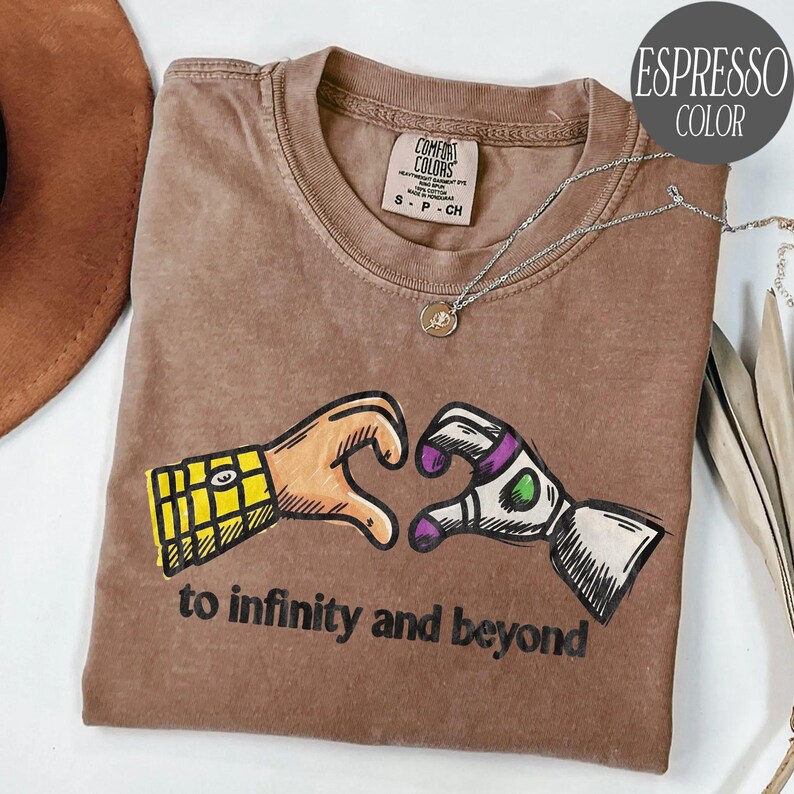 May include: An espresso-colored t-shirt featuring a graphic of two hands forming a heart shape, one with a yellow plaid pattern and the other with white, purple, and green details. The text "to infinity and beyond" is printed in black below.