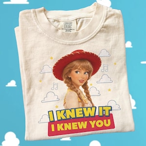 May include: A cream-colored t-shirt featuring a graphic of a character with blonde braided hair wearing a red cowboy hat. The design includes small clouds, musical notes, and yellow stars. Below the character, the text reads I KNEW IT I KNEW YOU.