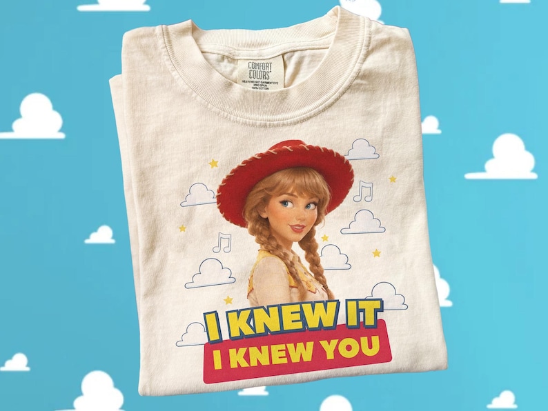 May include: A cream-colored t-shirt featuring a graphic of a character with blonde braided hair wearing a red cowboy hat. The design includes small clouds, musical notes, and yellow stars. Below the character, the text reads I KNEW IT I KNEW YOU.