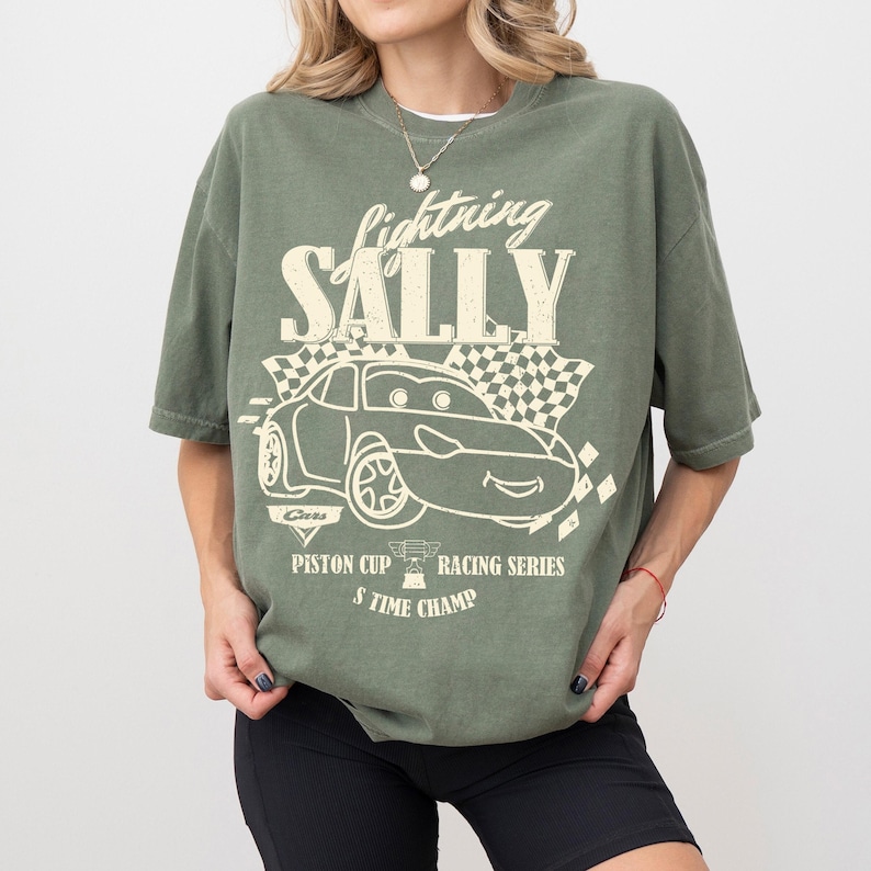 May include: An olive green t-shirt with a cartoon car graphic and the words "Lightning Sally". The design includes a checkered flag and the text "Piston Cup Racing Series" and "8 Time Champ".