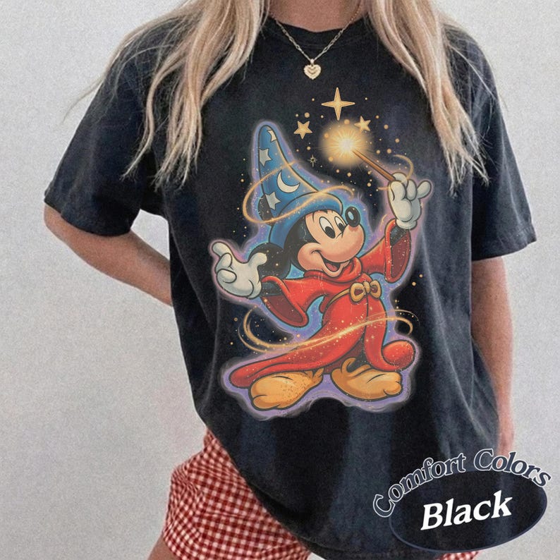 Pu&ograve; includere: T-shirt nero con un disegno di Topolino dei cartoni animati nei panni di un mago, che tiene in mano una bacchetta magica luminosa. Il design include stelle e vortici, con il testo "Comfort Colors Black". La maglietta ha maniche corte.