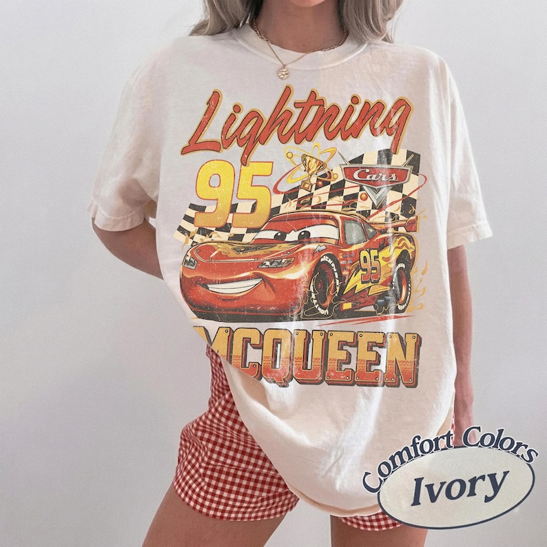 May include: Ivory-colored t-shirt featuring a graphic of Lightning McQueen from the Cars movie. The design includes the text "Lightning 95 McQueen" in red and yellow, with a checkered flag and trophy.