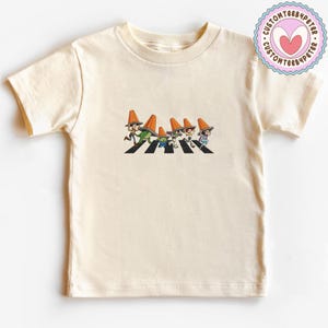 Puede incluir: Camiseta de color crema con una ilustraci&oacute;n de dibujos animados de personajes cruzando un paso de cebra, cada uno con un sombrero de cono naranja. El dise&ntilde;o es una parodia de una famosa portada de &aacute;lbum. Un logotipo circular est&aacute; en la esquina superior derecha.