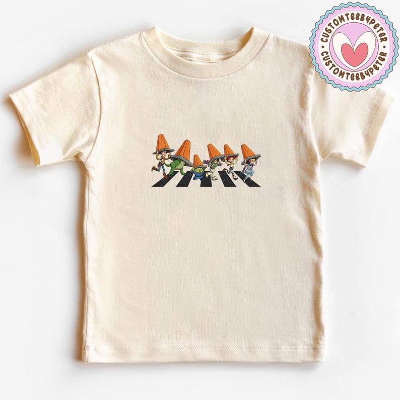 Puede incluir: Camiseta de color crema con una ilustraci&oacute;n de dibujos animados de personajes cruzando un paso de cebra, cada uno con un sombrero de cono naranja. El dise&ntilde;o es una parodia de una famosa portada de &aacute;lbum. Un logotipo circular est&aacute; en la esquina superior derecha.