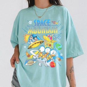 Pu&ograve; includere: T-shirt azzurra con una grafica colorata di personaggi Disney nello spazio. Il design include le parole "SPACE MOUNTAIN" in un font retr&ograve;, insieme a immagini di Topolino, Paperino e altri personaggi in astronavi e pianeti.