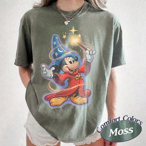 Pu&ograve; includere: T-shirt verde muschio con una grafica di Topolino come mago, che tiene in mano una bacchetta con una stella luminosa. Il design include un cappello da mago blu e vesti rosse. La maglietta &egrave; etichettata "Comfort Colors Moss".