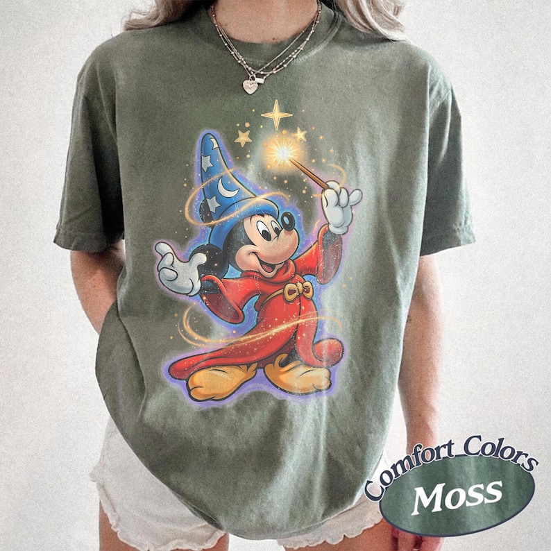 Pu&ograve; includere: T-shirt verde muschio con una grafica di Topolino come mago, che tiene in mano una bacchetta con una stella luminosa. Il design include un cappello da mago blu e vesti rosse. La maglietta &egrave; etichettata "Comfort Colors Moss".