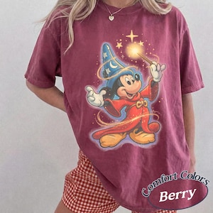 Pu&ograve; includere: T-shirt color bacca con una grafica di Topolino come mago, che tiene in mano una bacchetta luminosa. Mickey indossa un cappello da mago blu e vesti rosse. La maglietta ha le parole "Comfort Colors" e "Berry" stampate.