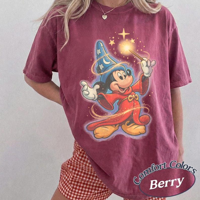 Pu&ograve; includere: T-shirt color bacca con una grafica di Topolino come mago, che tiene in mano una bacchetta luminosa. Mickey indossa un cappello da mago blu e vesti rosse. La maglietta ha le parole "Comfort Colors" e "Berry" stampate.