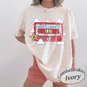 May include: An ivory t-shirt featuring a graphic of a red cassette tape with the text I KNEW IT, I KNEW YOU and TS. The design includes a yellow star with the number 13, white clouds, and musical notes surrounding the cassette tape illustration.