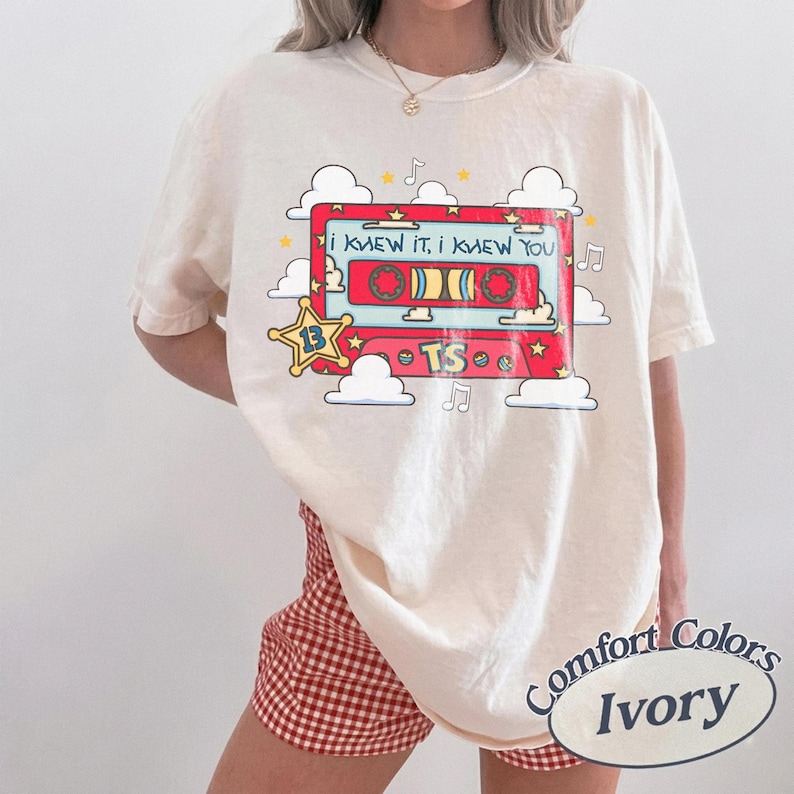 May include: An ivory t-shirt featuring a graphic of a red cassette tape with the text I KNEW IT, I KNEW YOU and TS. The design includes a yellow star with the number 13, white clouds, and musical notes surrounding the cassette tape illustration.