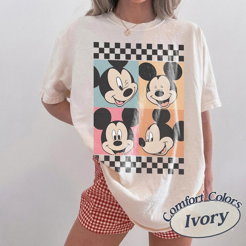 May include: Ivory-colored t-shirt featuring four panels with Mickey Mouse faces in different expressions. The panels are set against pastel backgrounds and framed by a black and white checkered pattern. The shirt has the words "Comfort Colors" and "Ivory" printed on it.