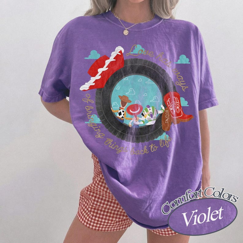May include: A violet short-sleeve t-shirt featuring a graphic of a black tire framing a blue sky with clouds. Inside the tire are cartoon characters, a red hat, and a red cowboy boot. The text reads Love has ways of bringing things back to life.