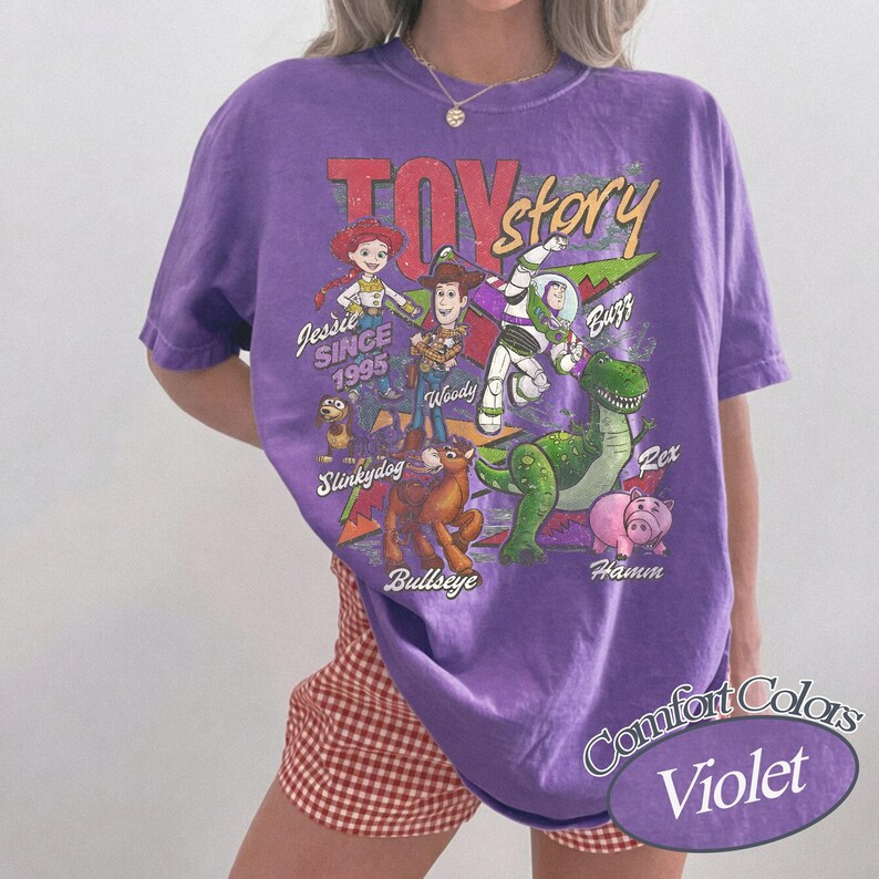 May include: A purple short-sleeve t-shirt featuring a graphic print of Toy Story characters including Jessie, Woody, Buzz Lightyear, Slinky Dog, Bullseye, Rex, and Hamm. The text reads Toy Story, Since 1995, and the names of the characters.