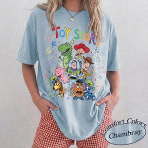 May include: Light blue t-shirt featuring a colorful graphic of Toy Story characters, including Woody, Buzz Lightyear, and Rex. The words "Toy Story" are written in a playful font above the characters. The shirt is a Comfort Colors Chambray.