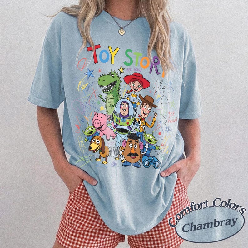 May include: Light blue t-shirt featuring a colorful graphic of Toy Story characters, including Woody, Buzz Lightyear, and Rex. The words "Toy Story" are written in a playful font above the characters. The shirt is a Comfort Colors Chambray.