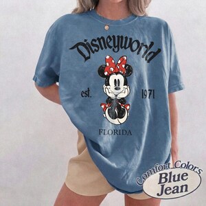 May include: Blue t-shirt with a graphic of Minnie Mouse wearing a red bow and the text "Disneyworld est. 1971 FLORIDA". The shirt is labeled "Comfort Colors Blue Jean".