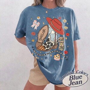 K&ouml;nnte beinhalten: Ein blaues Kurzarm-T-Shirt mit einem Grafikdruck, der einen roten Cowboyhut, einen Stiefel mit Kuhmuster und dem Namen Taylor, einen Stapel Bl&ouml;cke mit den Buchstaben T S, eine Schleife, Blumen, Sterne und eine Perlenkette zeigt.