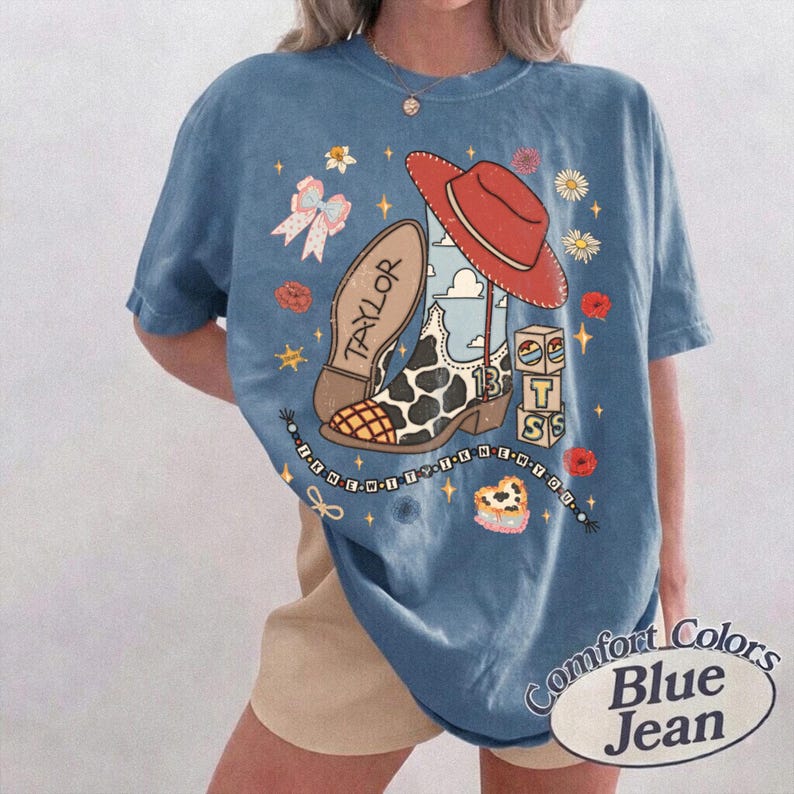K&ouml;nnte beinhalten: Ein blaues Kurzarm-T-Shirt mit einem Grafikdruck, der einen roten Cowboyhut, einen Stiefel mit Kuhmuster und dem Namen Taylor, einen Stapel Bl&ouml;cke mit den Buchstaben T S, eine Schleife, Blumen, Sterne und eine Perlenkette zeigt.