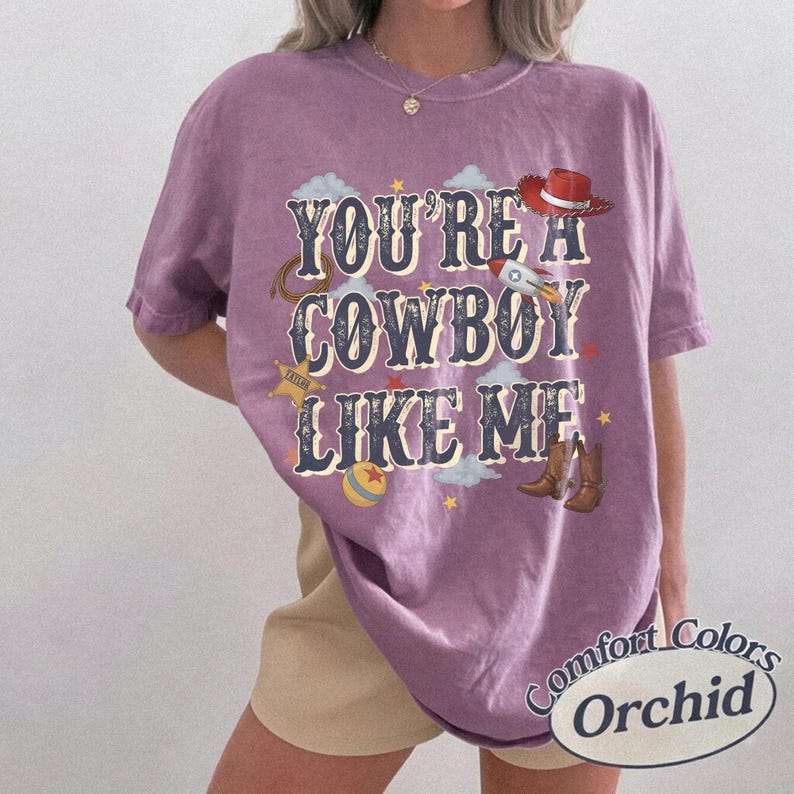 May include: A purple short-sleeve t-shirt featuring the text "YOU'RE A COWBOY LIKE ME" in a dark blue font. The graphic includes illustrations of a red cowboy hat, a rocket, a lasso, a sheriff star, a ball, and brown cowboy boots with clouds.