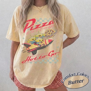 Pu&ograve; includere: T-shirt color burro con grafica retr&ograve; di tre alieni verdi su un razzo, una pizza e un pianeta. Il testo recita "Pizza H-O-T-T-O-GO" e "You can take me Hot to Go".