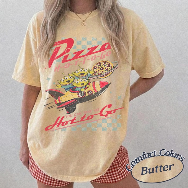 Pu&ograve; includere: T-shirt color burro con grafica retr&ograve; di tre alieni verdi su un razzo, una pizza e un pianeta. Il testo recita "Pizza H-O-T-T-O-GO" e "You can take me Hot to Go".