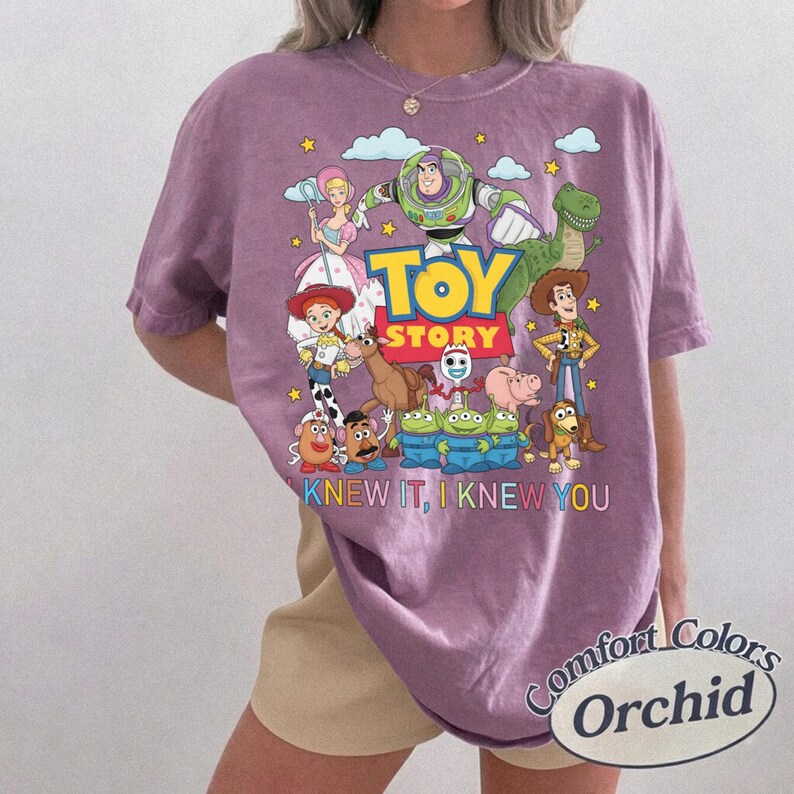 May include: A purple short-sleeve t-shirt featuring a graphic print of Toy Story characters including Buzz Lightyear, Woody, Jessie, and others. The text "TOY STORY" is centered above the phrase "I KNEW IT, I KNEW YOU" in colorful lettering.