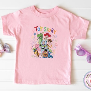 May include: A light pink t-shirt featuring a colorful graphic of characters from the movie "Toy Story." The design includes the words "Toy Story" in a playful font, along with images of Woody, Buzz Lightyear, and other characters.
