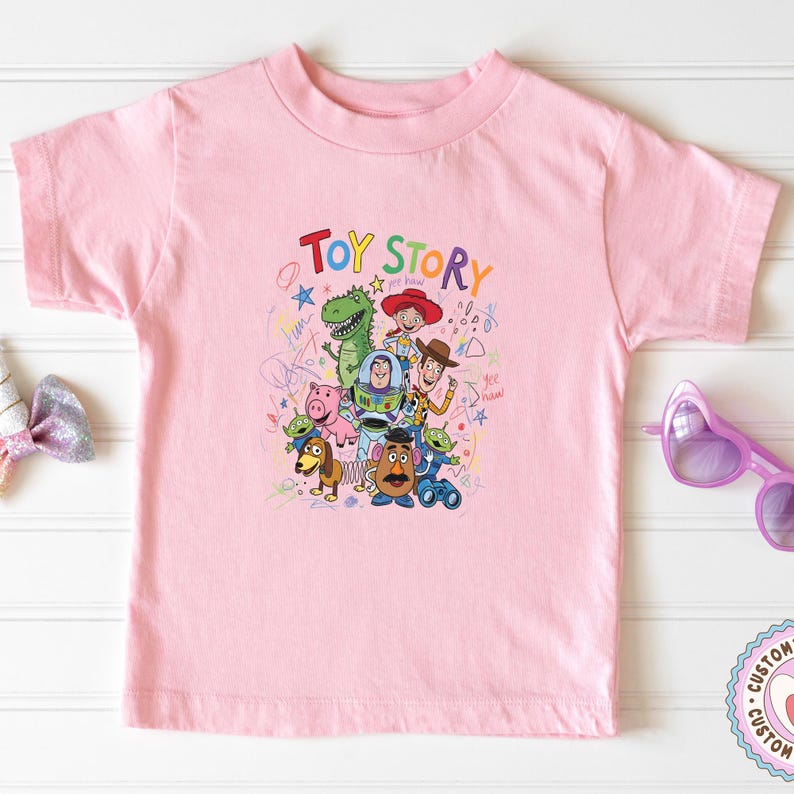 May include: A light pink t-shirt featuring a colorful graphic of characters from the movie "Toy Story." The design includes the words "Toy Story" in a playful font, along with images of Woody, Buzz Lightyear, and other characters.
