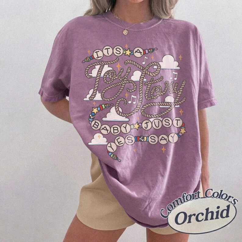 May include: An orchid-colored short-sleeve t-shirt featuring a graphic print with the text "ITS A TOY STORY BABY JUST YES SAY". The design includes rope-style lettering, colorful beads, small stars, musical notes, and white clouds on the front.