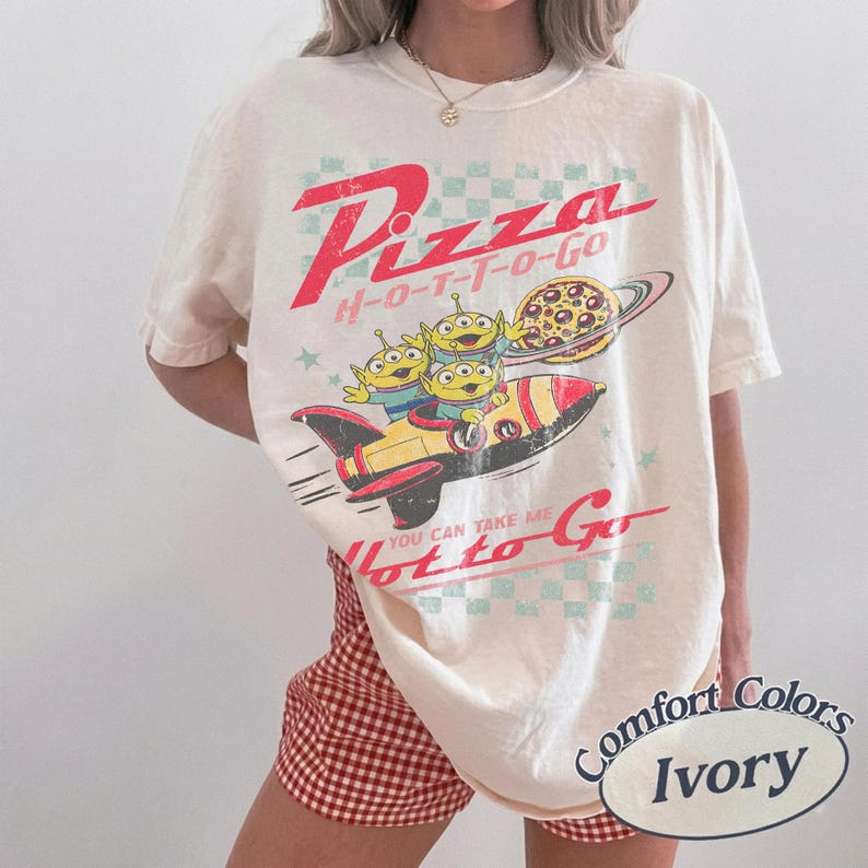 Pu&ograve; includere: T-shirt color avorio con una grafica di tre alieni verdi su una navicella spaziale, un pianeta pizza e le parole "Pizza H-O-T-T-O-Go". La maglietta ha anche il testo "You can take me Hot to Go" e "Comfort Colors Ivory".