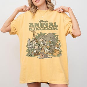 Puede incluir: Camiseta amarilla mostaza con el texto "Disney's Animal Kingdom" en verde. El gr&aacute;fico representa personajes de Disney como Mickey Mouse, Donald Duck y varios animales, incluyendo una jirafa y un tigre.