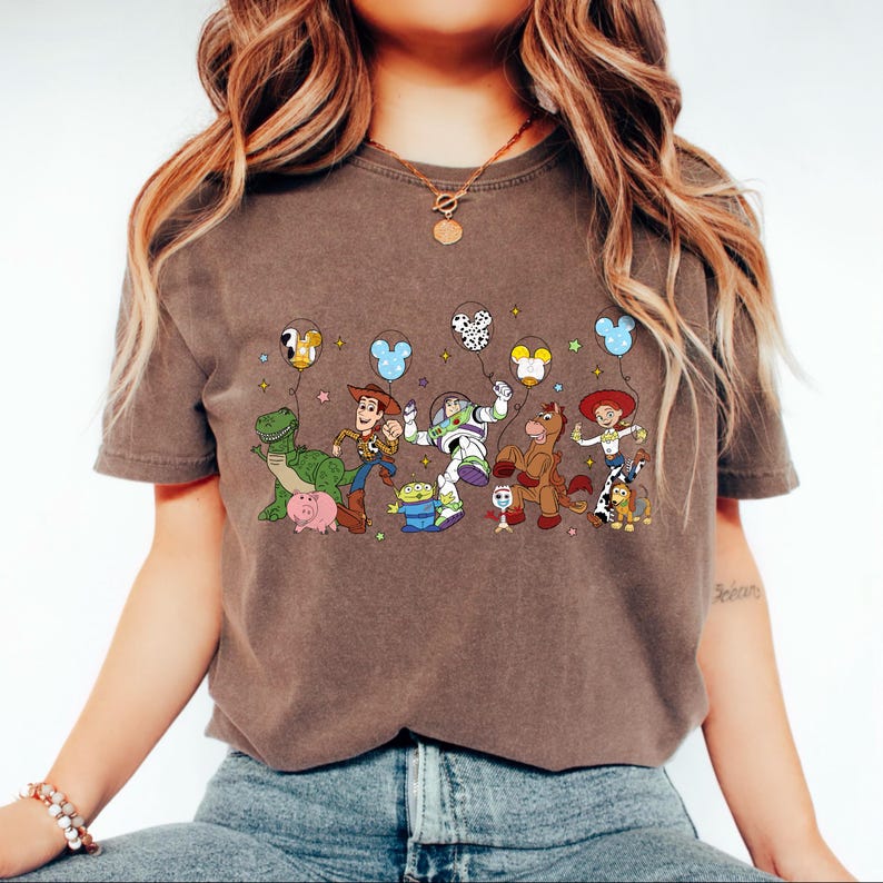 May include: Brown t-shirt featuring a colorful graphic of animated characters, including a green dinosaur, a cowboy, and a space ranger, holding balloons. The design includes various characters from a popular animated movie. The shirt is made of a soft material.