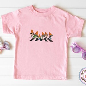 Puede incluir: Camiseta rosa claro con un gr&aacute;fico de dibujos animados de personajes cruzando una calle. Los personajes llevan sombreros de cono naranja. La camiseta es de cuello redondo y manga corta.