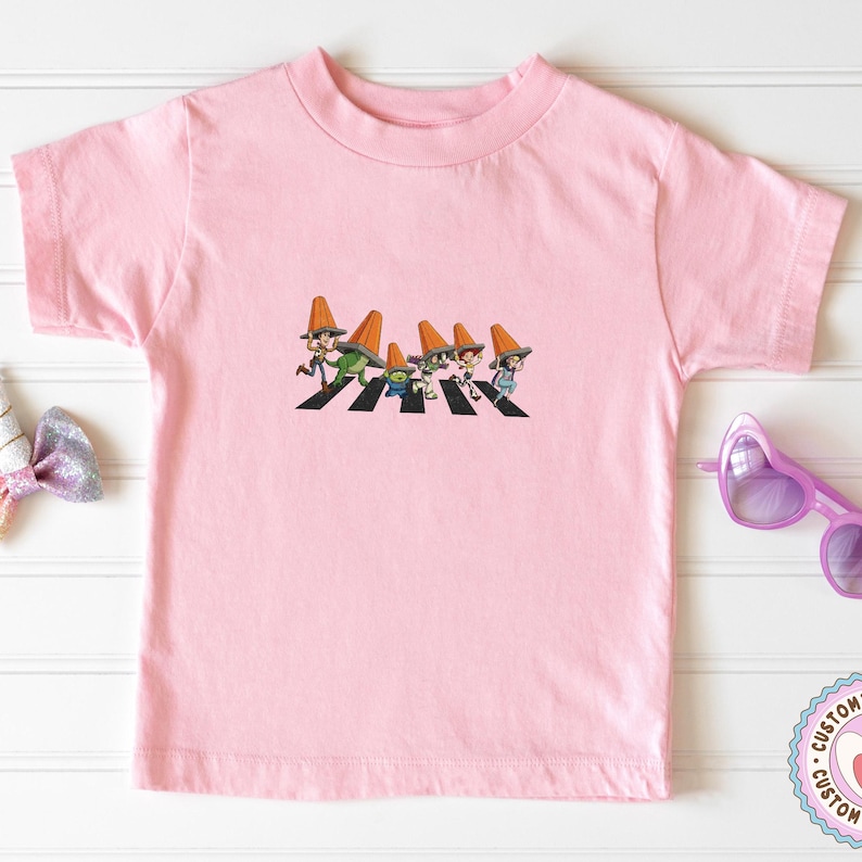 Puede incluir: Camiseta rosa claro con un gr&aacute;fico de dibujos animados de personajes cruzando una calle. Los personajes llevan sombreros de cono naranja. La camiseta es de cuello redondo y manga corta.