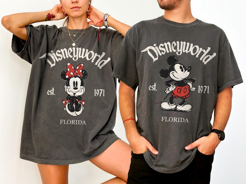 May include: Two charcoal gray t-shirts featuring Disney characters. One shirt displays Minnie Mouse with a red bow, and the other shows Mickey Mouse. Both shirts have "Disneyworld est. 1971 FLORIDA" printed on them.