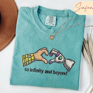 May include: A seafoam green t-shirt featuring a graphic of two hands forming a heart shape, one resembling a yellow plaid sleeve and the other a white and purple space suit sleeve. The text "to infinity and beyond" is printed below the graphic.