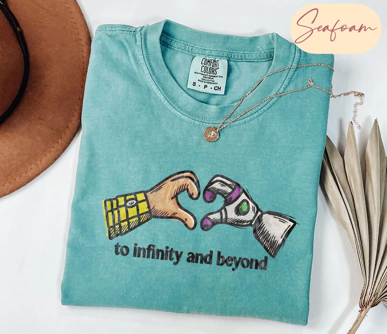 May include: A seafoam green t-shirt featuring a graphic of two hands forming a heart shape, one resembling a yellow plaid sleeve and the other a white and purple space suit sleeve. The text "to infinity and beyond" is printed below the graphic.