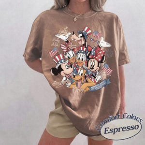 Puede incluir: Camiseta marr&oacute;n Comfort Colors Espresso con un dise&ntilde;o de los personajes de Disney Mickey Mouse, Minnie Mouse, Donald, Daisy, Goofy y Pluto, todos con sombreros patri&oacute;ticos y banderas americanas. La camiseta conmemora el 250 aniversario de EE. UU.