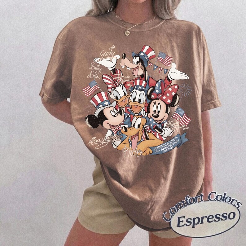 Puede incluir: Camiseta marr&oacute;n Comfort Colors Espresso con un dise&ntilde;o de los personajes de Disney Mickey Mouse, Minnie Mouse, Donald, Daisy, Goofy y Pluto, todos con sombreros patri&oacute;ticos y banderas americanas. La camiseta conmemora el 250 aniversario de EE. UU.