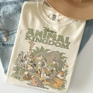 Puede incluir: Camiseta color crema con el texto "Disney's Animal Kingdom" en verde. La camiseta muestra varios personajes de Disney, incluyendo Mickey Mouse y Donald Duck, sobre un fondo de jungla. La camiseta est&aacute; doblada sobre unos vaqueros azules.