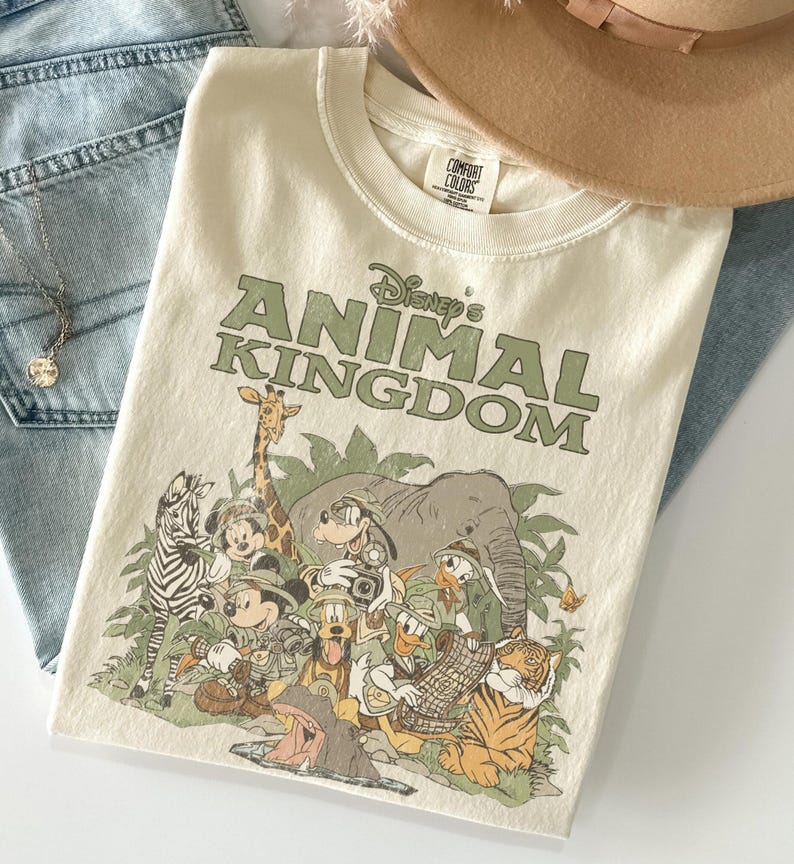Puede incluir: Camiseta color crema con el texto "Disney's Animal Kingdom" en verde. La camiseta muestra varios personajes de Disney, incluyendo Mickey Mouse y Donald Duck, sobre un fondo de jungla. La camiseta est&aacute; doblada sobre unos vaqueros azules.