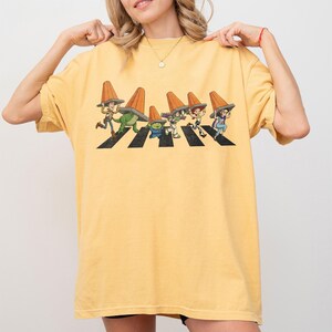 Puede incluir: Camiseta amarilla mostaza con una ilustraci&oacute;n de dibujos animados de personajes cruzando un paso de cebra, cada uno con un cono de tr&aacute;fico naranja. El dise&ntilde;o es una parodia de la ic&oacute;nica portada del &aacute;lbum Abbey Road.