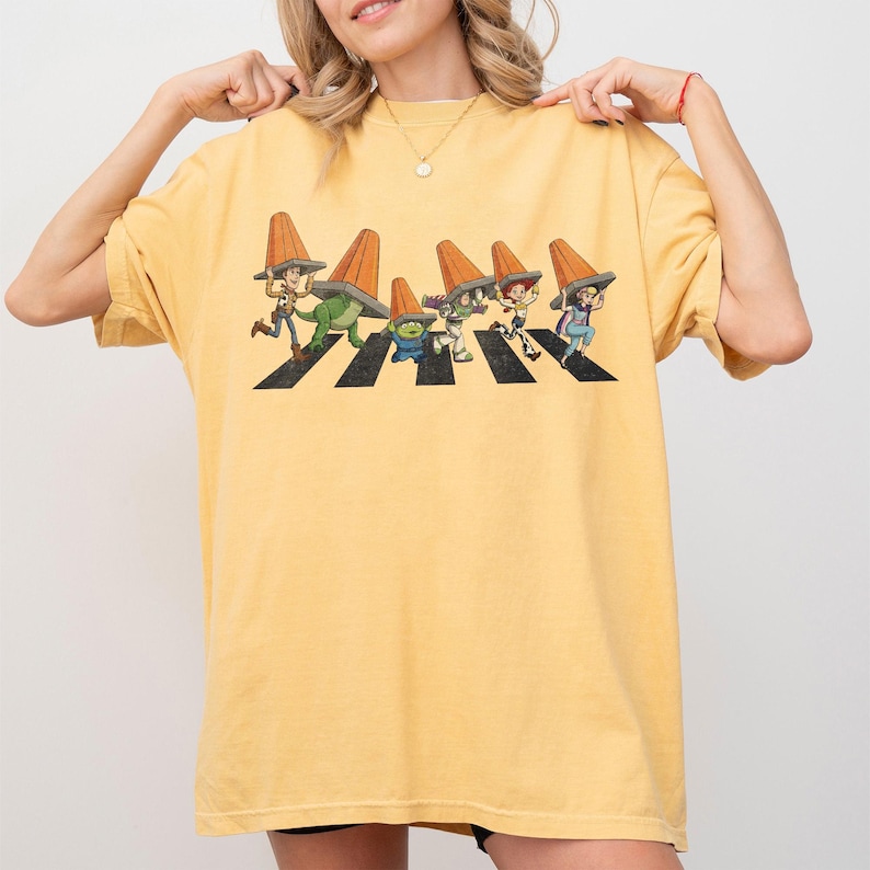 Puede incluir: Camiseta amarilla mostaza con una ilustraci&oacute;n de dibujos animados de personajes cruzando un paso de cebra, cada uno con un cono de tr&aacute;fico naranja. El dise&ntilde;o es una parodia de la ic&oacute;nica portada del &aacute;lbum Abbey Road.