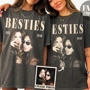 以下が含まれることがあります： クリーム色のセリフ体で「BESTIES EST. 2018」とプリントされたチャコールグレーの半袖Tシャツ。テキストの下には、ワイングラスを持った2人の人物のモノクロ写真がプリントされており、そのうち1人はサングラスをかけている。